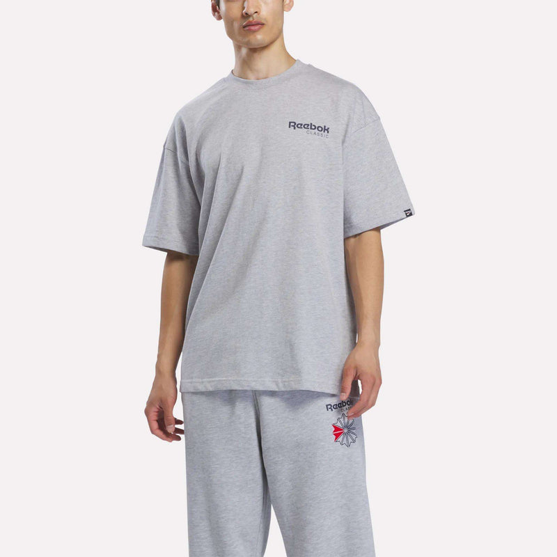 Wearing a Men’s Relaxed Logo T-Shirt by Reebok in light gray, along with matching logo sweatpants, a person stands against a plain white background.