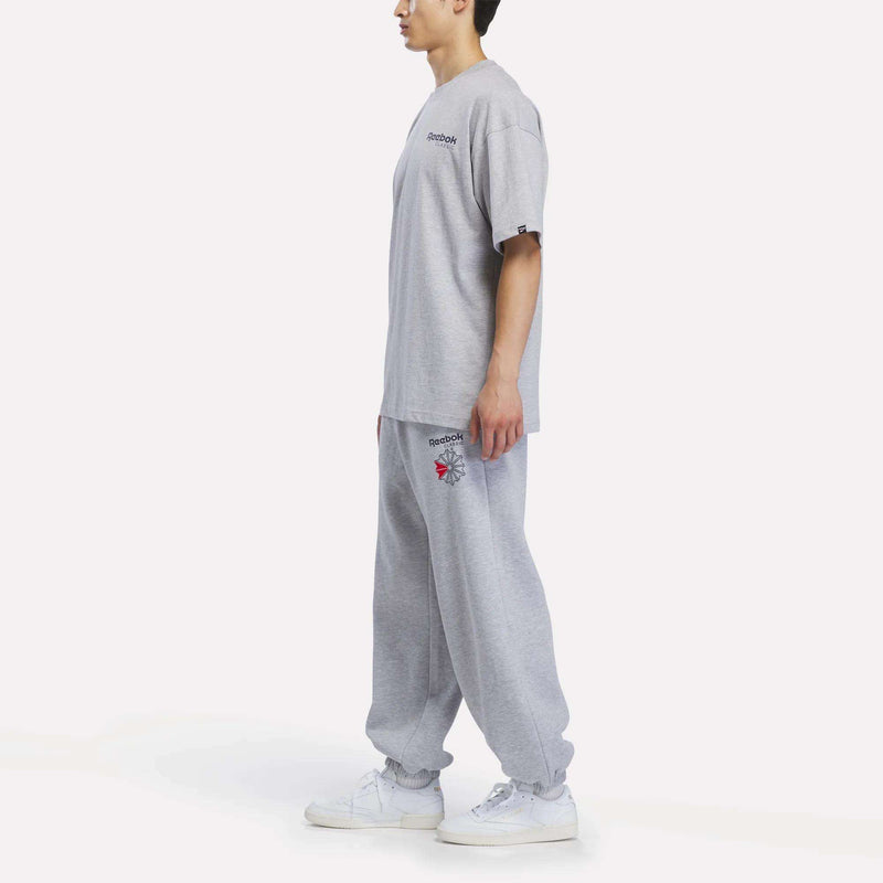 A person stands sideways wearing a Reebok Men’s Relaxed Logo T-Shirt, crafted from soft cotton, paired with matching gray sweatpants and white sneakers. The background is plain white.