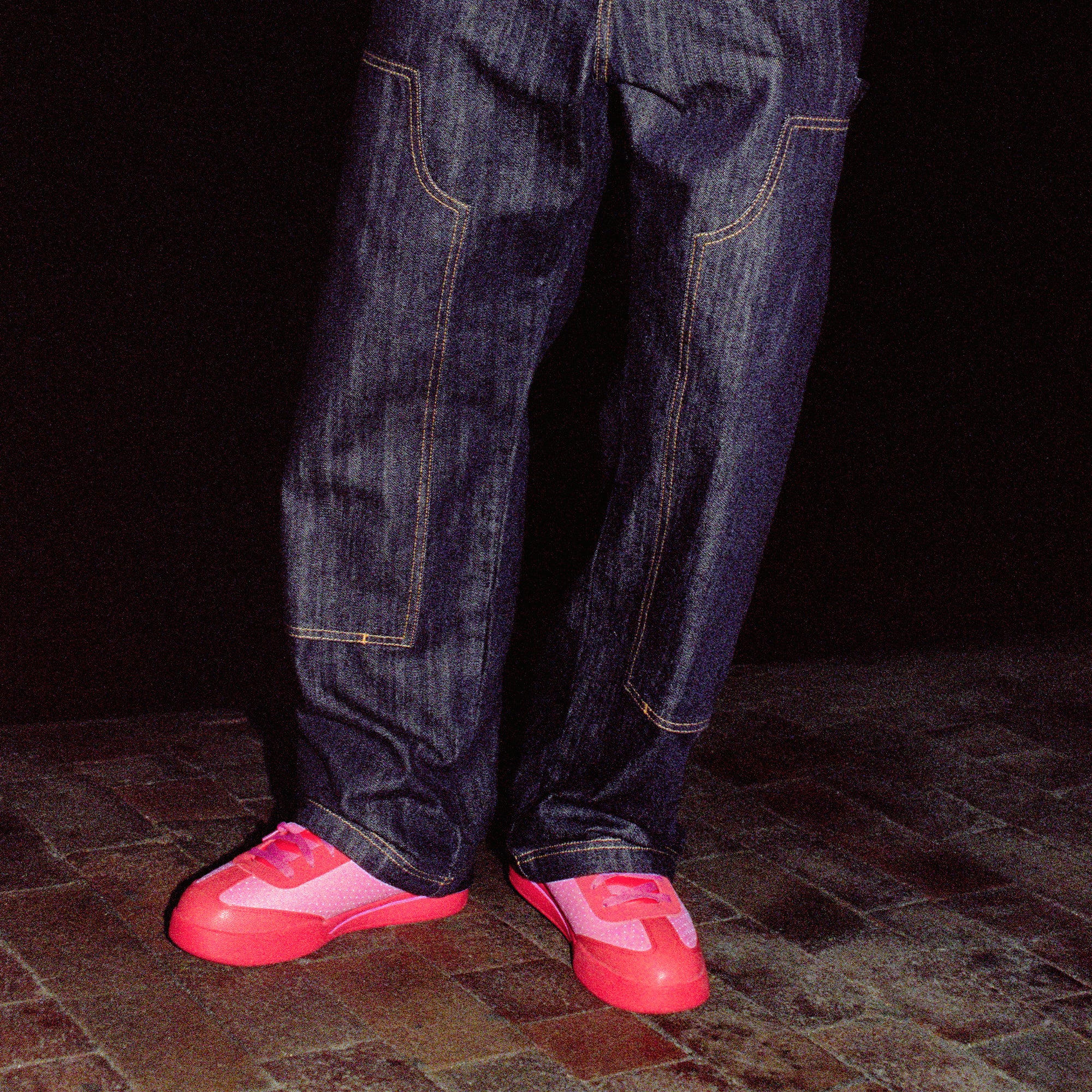 A person stands on a brick surface at night in wide-leg dark denim jeans with visible stitching, wearing bright pink Reebok x BBC Board Flip Skate Shoes featuring light pink and purple details. The background is mostly dark.
