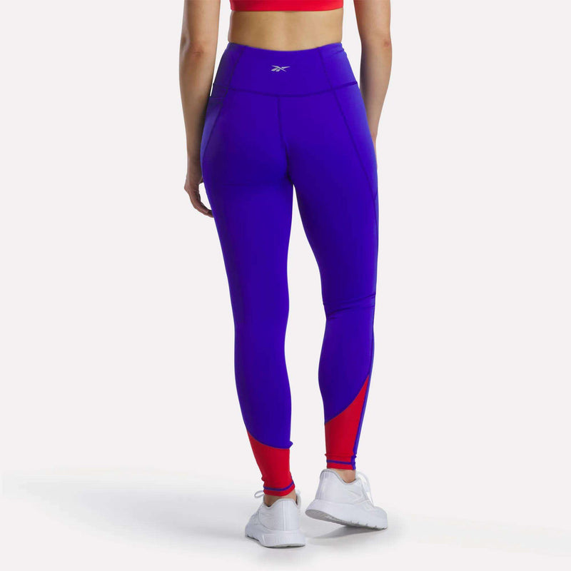 A person wearing a red sports bra, white sneakers, and Reebok Women's Running Leggings—high-waisted purple leggings featuring mesh panels and bold red accents near the ankles—stands with their back to the camera.