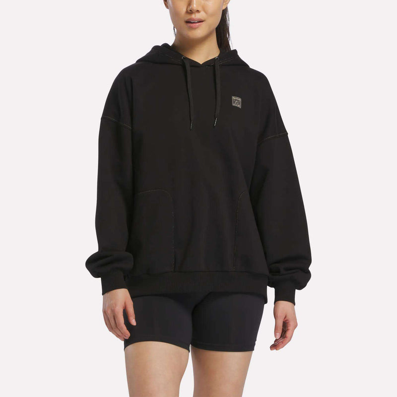 A woman stands against a plain white background, facing slightly to the side. She is wearing the Reebok Women's Relaxed Fleece Hoodie in black, paired with black shorts. Her long dark hair is pulled back, and her hands rest relaxed by her sides.