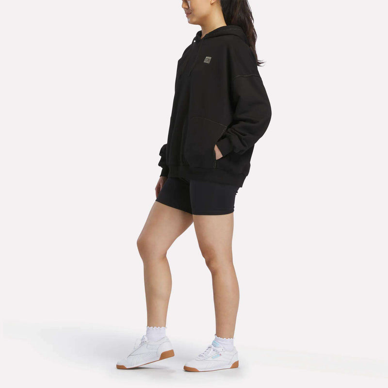 A person stands sideways against a plain white background wearing the Reebok Women's Relaxed Fleece Hoodie in black, paired with black shorts, white socks, and white sneakers.
