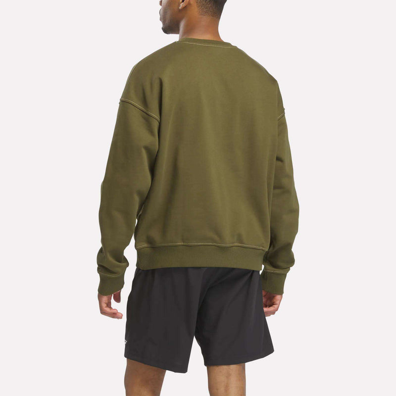 A man faces away, wearing a relaxed fit Reebok Men's Patch Logo Crewneck Sweatshirt in olive green and black shorts, set against a plain white background.
