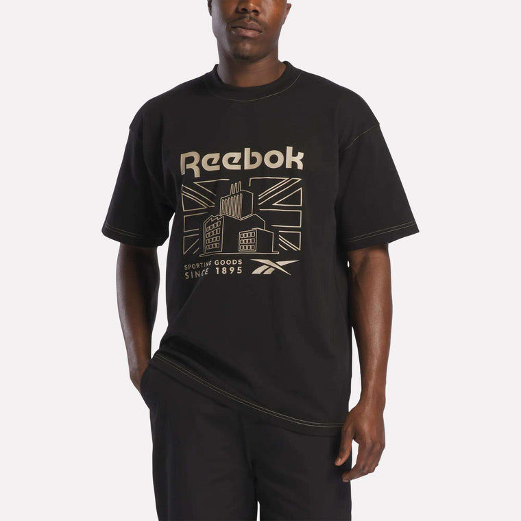 A person wearing the Reebok Men's Big Graphic T-Shirt in black, paired with black pants, stands against a plain white background.