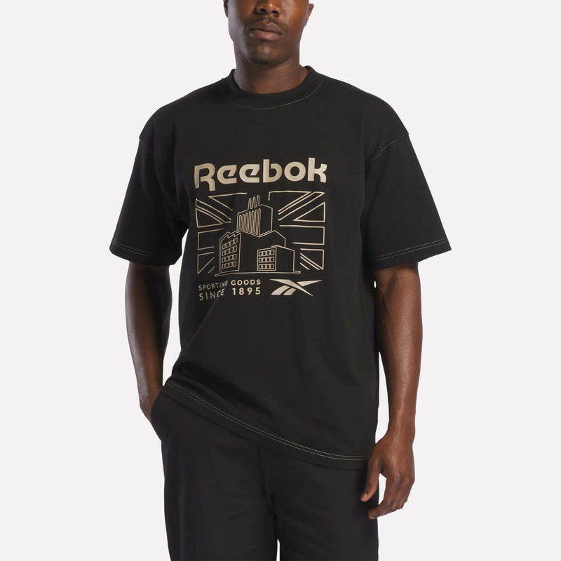 A person wearing the Reebok Men's Big Graphic T-Shirt in black, paired with black pants, stands against a plain white background.