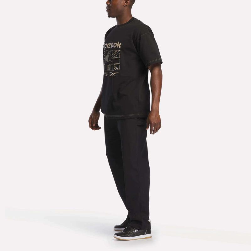 A person stands against a plain background, wearing the Reebok Men's Big Graphic T-Shirt in black with a crewneck design, paired with black pants and black Reebok sneakers. This cotton jersey tee features a large Reebok logo prominently displayed on the front.