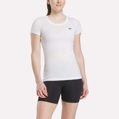 A woman wears the Women's Reebok T-Shirt, a fitted white tee with a small logo on the chest, paired with black shorts, standing against a plain light background.