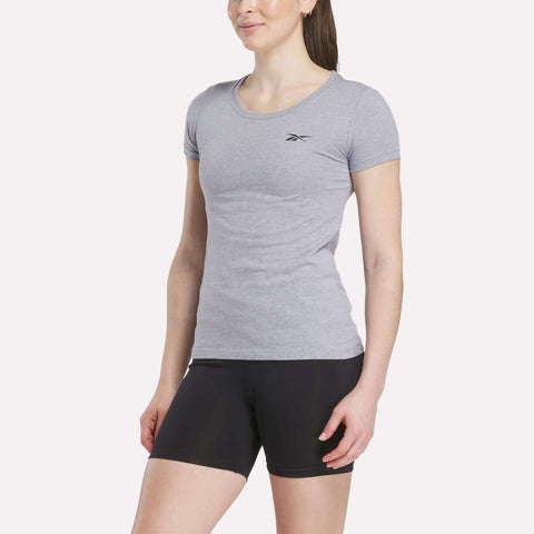 A woman stands against a plain light background wearing a fitted Women's Reebok T-Shirt in light gray with a small chest logo, paired with black shorts.