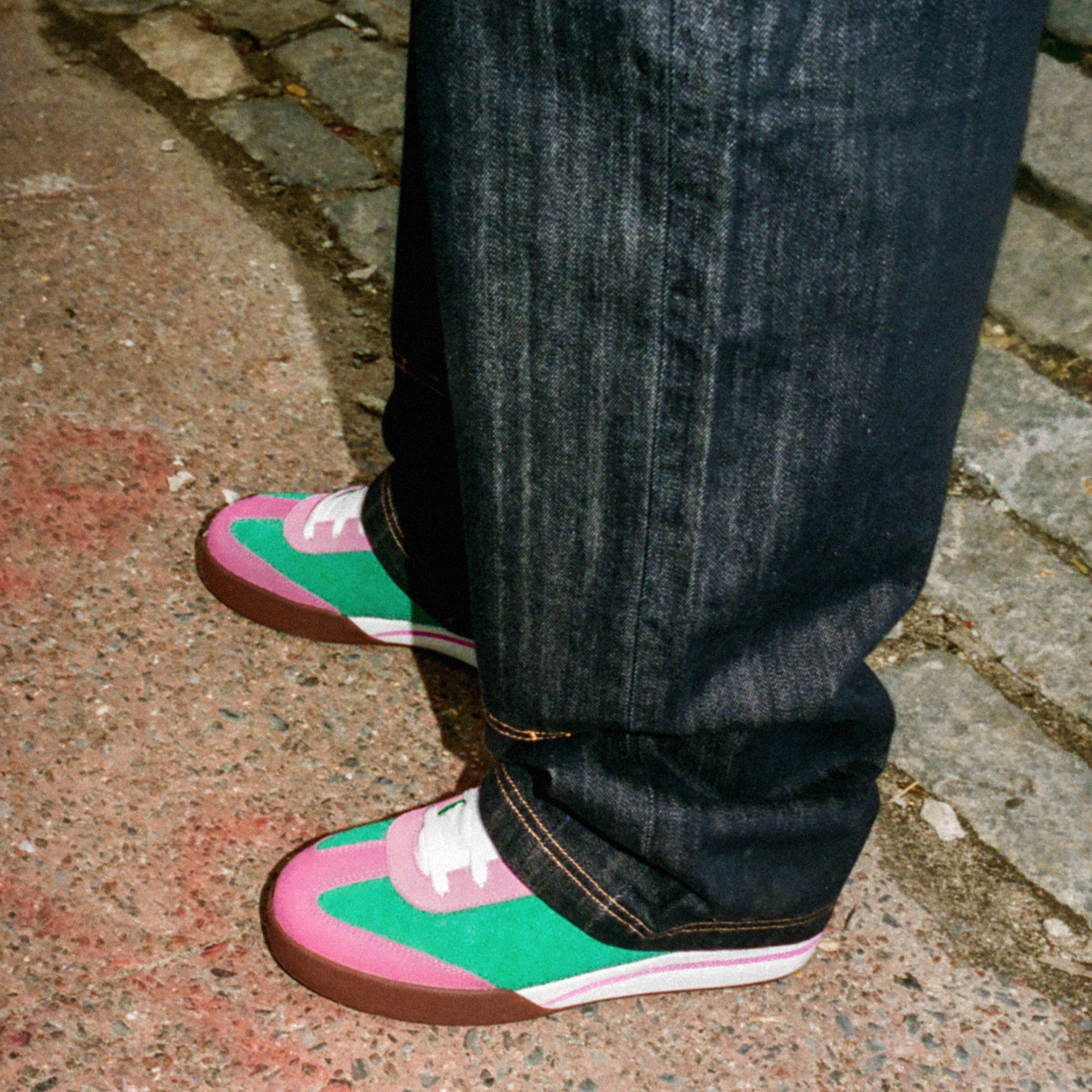A person on a sidewalk wears dark jeans and Reebok x BBC Board Flip Skate Shoes featuring pink, green, white, and brown suede accents. Cobblestone and pavement are visible underfoot.