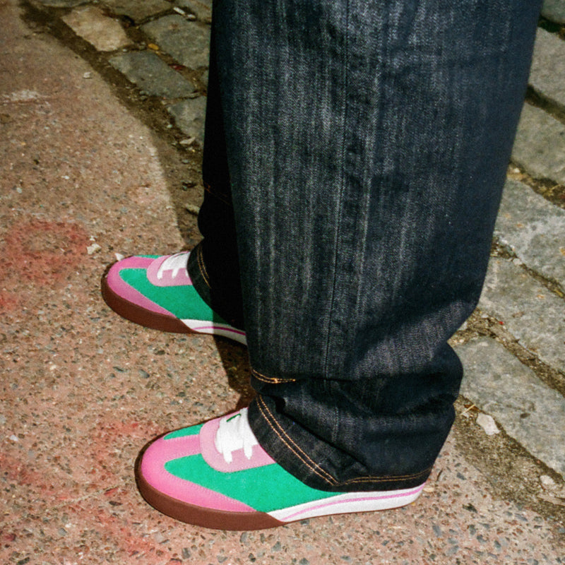 A person on a sidewalk wears dark jeans and Reebok x BBC Board Flip Skate Shoes featuring pink, green, white, and brown suede accents. Cobblestone and pavement are visible underfoot.