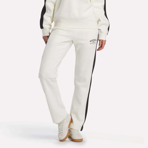 A person is wearing the Reebok Women's Fleece Straight Leg Pants in chalk, featuring black side stripes and a small logo on the upper thigh, paired with white sneakers. Their hands are at their sides against a plain white background.