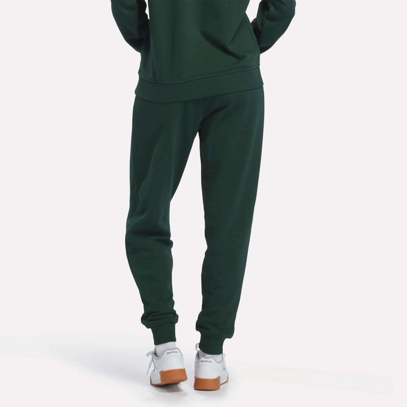 A person is shown from the waist down, wearing Reebok Women’s French Terry Joggers in dark green with a heritage-inspired design, paired with white Reebok sneakers featuring light brown soles, set against a white background.