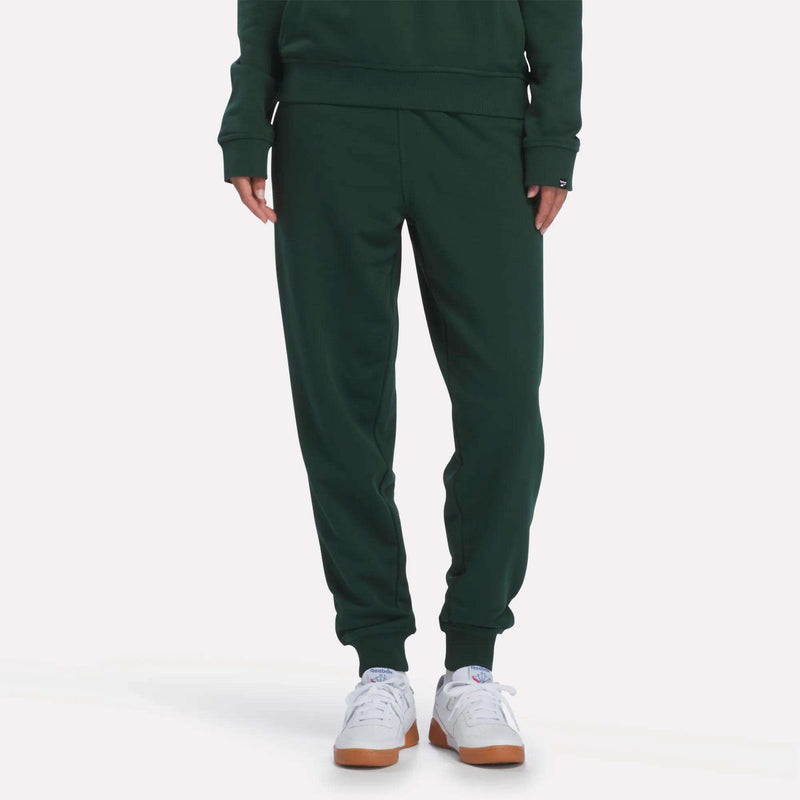 A woman is wearing Reebok Women’s French Terry Joggers in Heritage Green, paired with a coordinating sweatshirt and crisp white Reebok athletic shoes with gum soles, standing against a plain white background—a look that merges modern comfort with the timeless heritage style of Reebok.