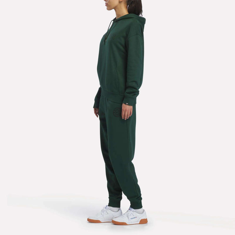 A woman stands sideways on a plain white background, wearing Reebok Women’s French Terry Joggers in dark green with a matching hoodie and white sneakers—showcasing a sporty heritage design.