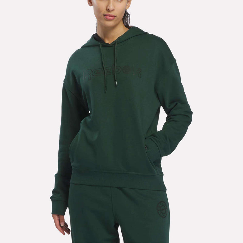 A woman wearing the Reebok Women’s French Terry Lawn Club Hoodie in Heritage Green stands with her hands in the pockets against a plain white background.