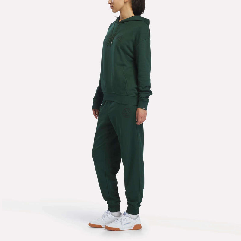 A person stands in a dark green Reebok Women’s French Terry Lawn Club Hoodie and matching joggers with white sneakers, facing slightly left, against a plain white background.