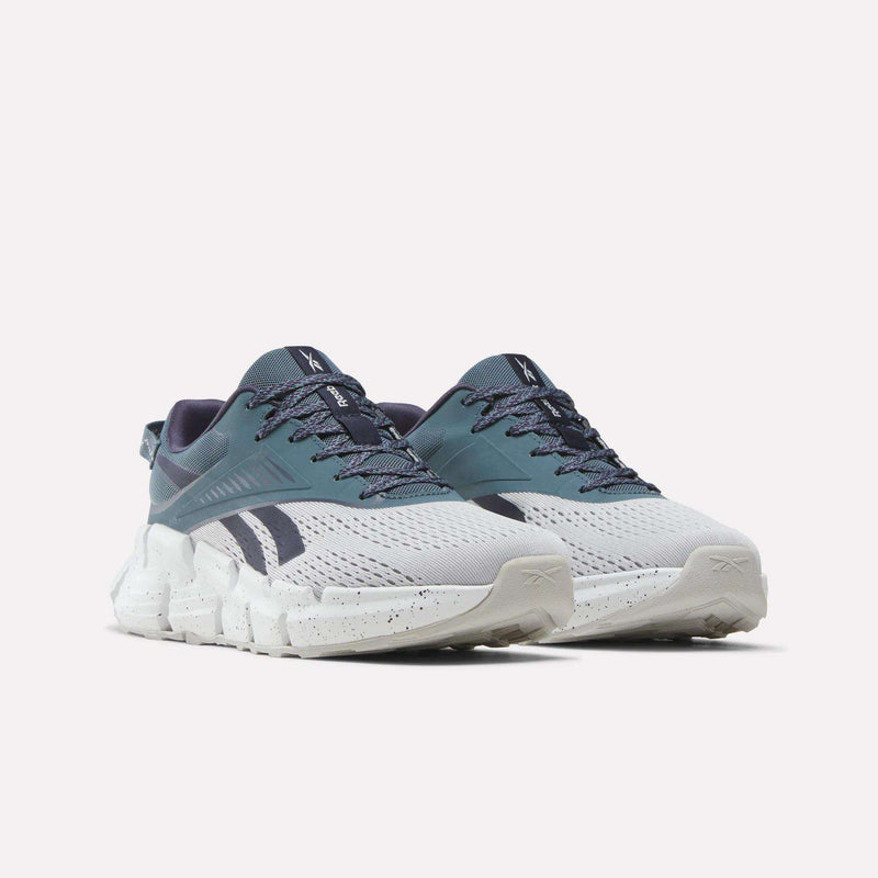 Pair of Reebok Zig Dynamica 6 Running Shoes for big kids in white and teal, featuring dark blue accents, chunky white soles with Zig technology, and patterned blue laces, displayed against a plain white background.