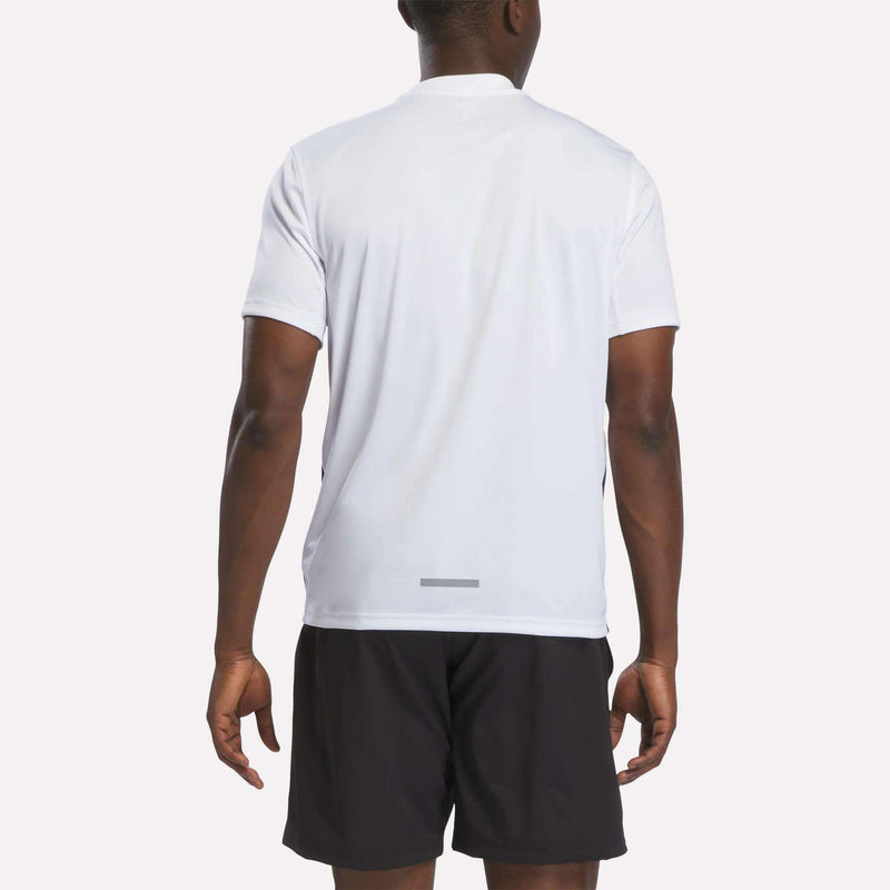 A man stands facing away from the camera, wearing a white Reebok Men’s Short Sleeve Vector T-Shirt and black shorts against a plain background.