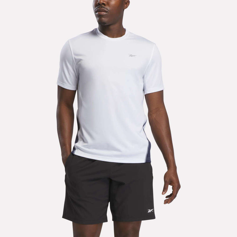 A man wearing a white Reebok Men’s Short Sleeve Vector T-Shirt in White/Vector Navy, featuring RBK-DRY technology and a small logo, along with black shorts with a small logo, stands against a plain background.
