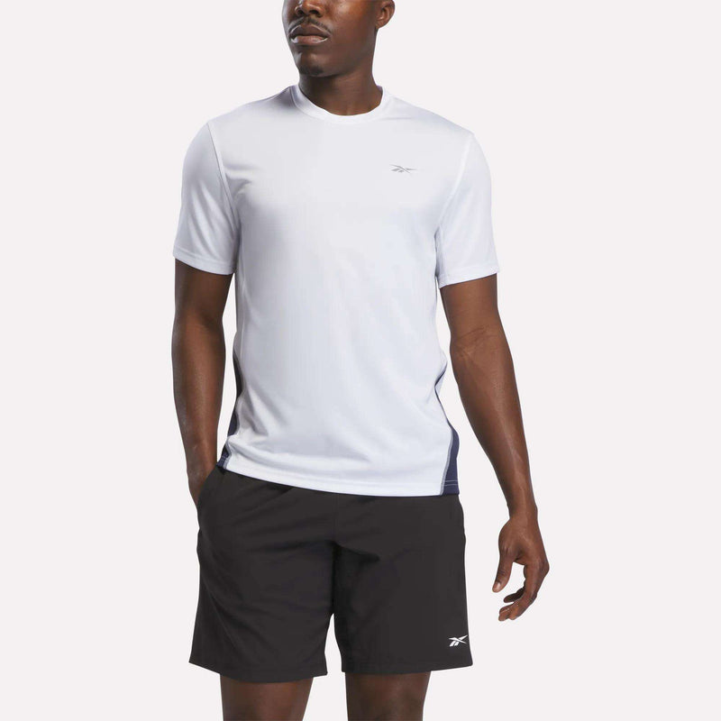 A man wearing a white Reebok Men’s Short Sleeve Vector T-Shirt in White/Vector Navy, featuring RBK-DRY technology and a small logo, along with black shorts with a small logo, stands against a plain background.