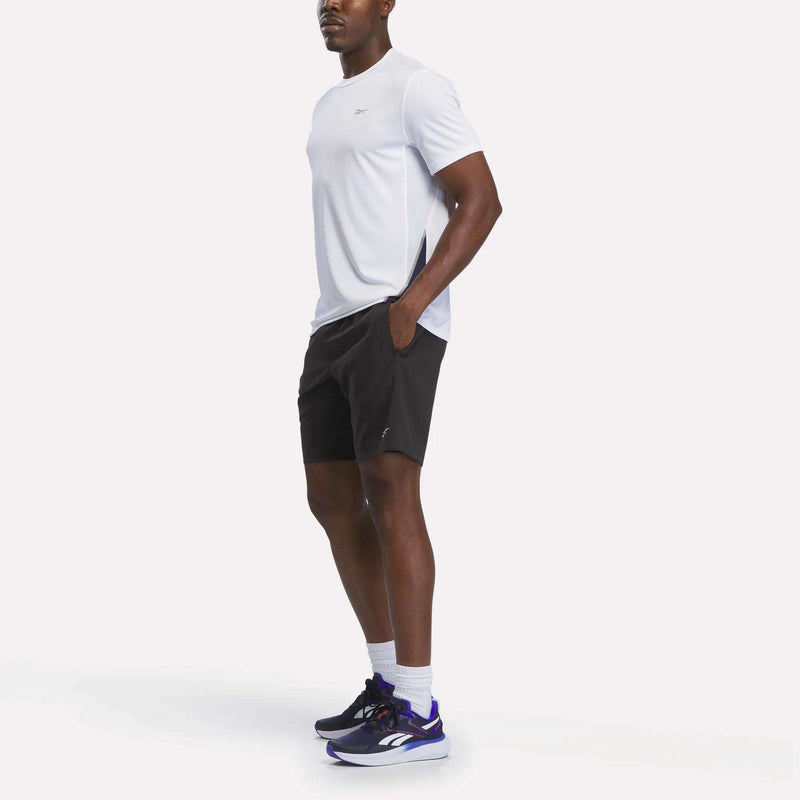 A man stands with his hands in his pockets, wearing the Reebok Men’s Short Sleeve Vector T-Shirt in white, which features RBK-DRY technology. He pairs it with black shorts, white socks, and black and purple sneakers against a plain white background.