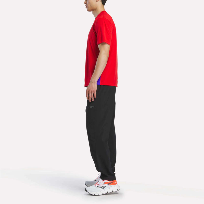 A man stands sideways against a plain white background, wearing a red Reebok Men's Short Sleeve Vector T-Shirt with RBK-DRY technology, black pants, and white athletic shoes with orange accents.