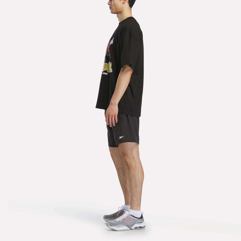 A person in profile wears a black Reebok Men's Basketball T-Shirt, black athletic shorts, and gray athletic shoes, standing against a plain white background.