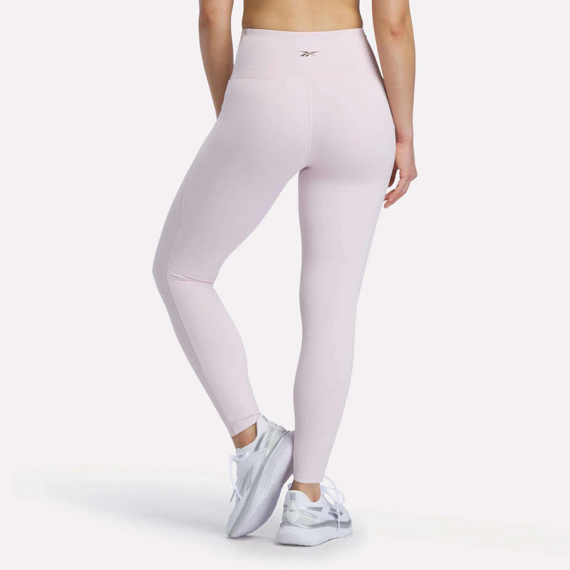 A person is wearing Reebok Women's High-Rise Leggings in light pink, which feature RBK-DRY technology, along with white athletic shoes, standing with their back to the camera against a plain white background.
