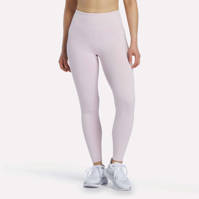 A woman wearing Reebok Women's High-Rise Leggings in Elemental Pink and white sneakers, standing against a plain white background. Her upper body and head are not visible.