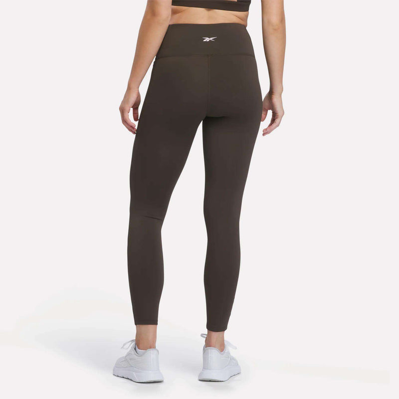 A person is wearing Reebok Women’s High-Rise Leggings in dark brown, featuring RBK-DRY fabric, paired with a matching sports bra and white sneakers, standing with their back to the camera against a plain white background.