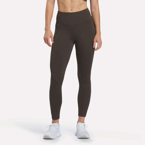 A person is shown from the neck down, standing against a plain white background and wearing Reebok Women’s High-Rise Leggings in Espresso along with white sneakers. The leggings are crafted with RBK-DRY performance fabric.