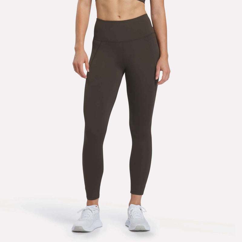 A person is shown from the neck down, standing against a plain white background and wearing Reebok Women’s High-Rise Leggings in Espresso along with white sneakers. The leggings are crafted with RBK-DRY performance fabric.