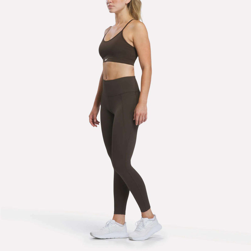 A woman stands in a relaxed pose against a plain white background, wearing Reebok Women’s High-Rise Leggings and a matching sports bra crafted from RBK-DRY performance fabric, paired with white sneakers.