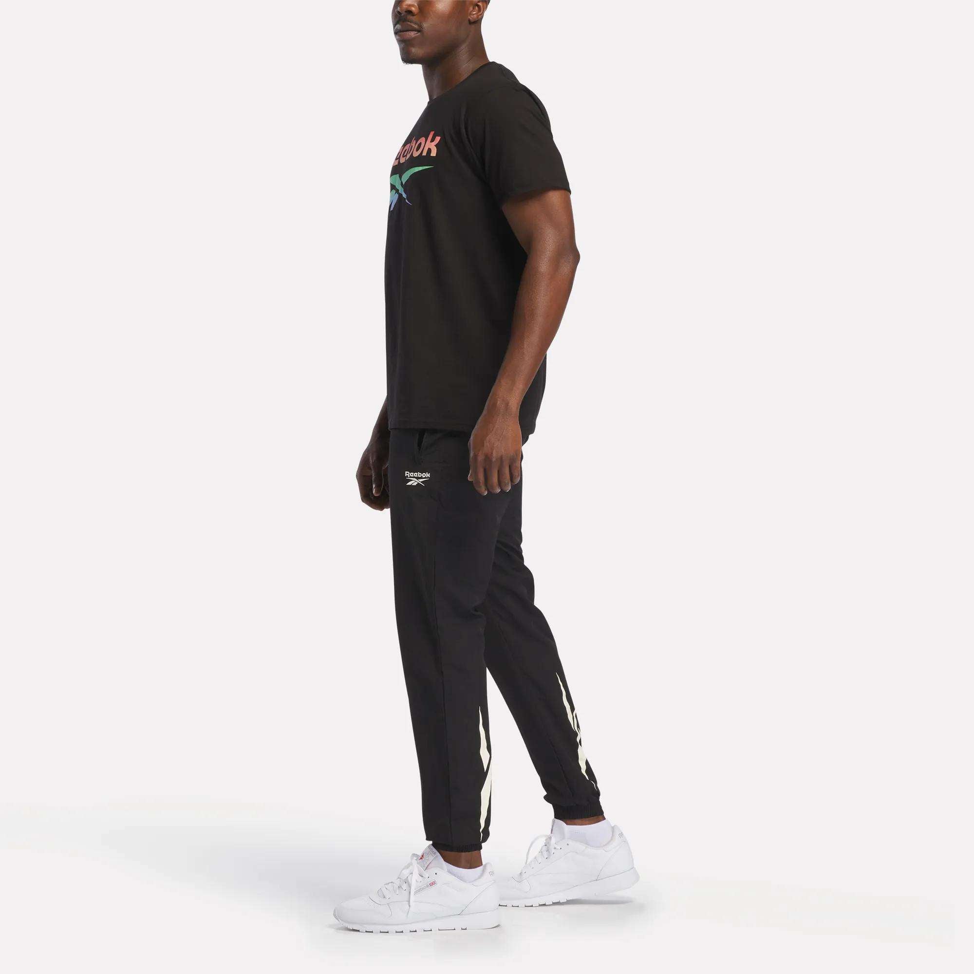 A man stands slightly sideways, wearing Reebok's Men's Vector Graphic Rainbow T-Shirt in black, paired with black joggers with white logo details and white sneakers, against a plain white background.