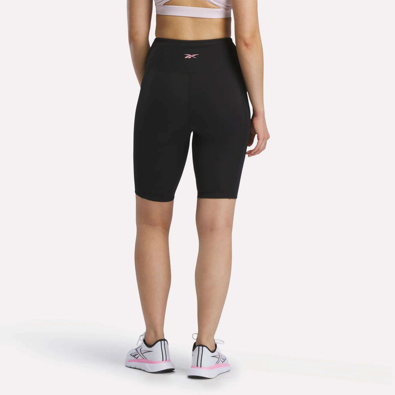 A person stands facing away against a plain white background, wearing a white sports bra, Reebok Women's Peached Biker Shorts 9" in black made from peached fabric, and white sneakers with pink accents, showing the back view of the outfit.