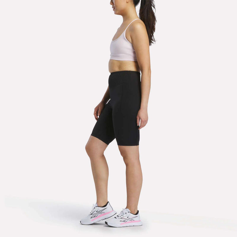 A woman stands in profile against a plain white background, wearing a light sports bra, Reebok Women's Peached Biker Shorts 9", and white athletic shoes with pink accents.