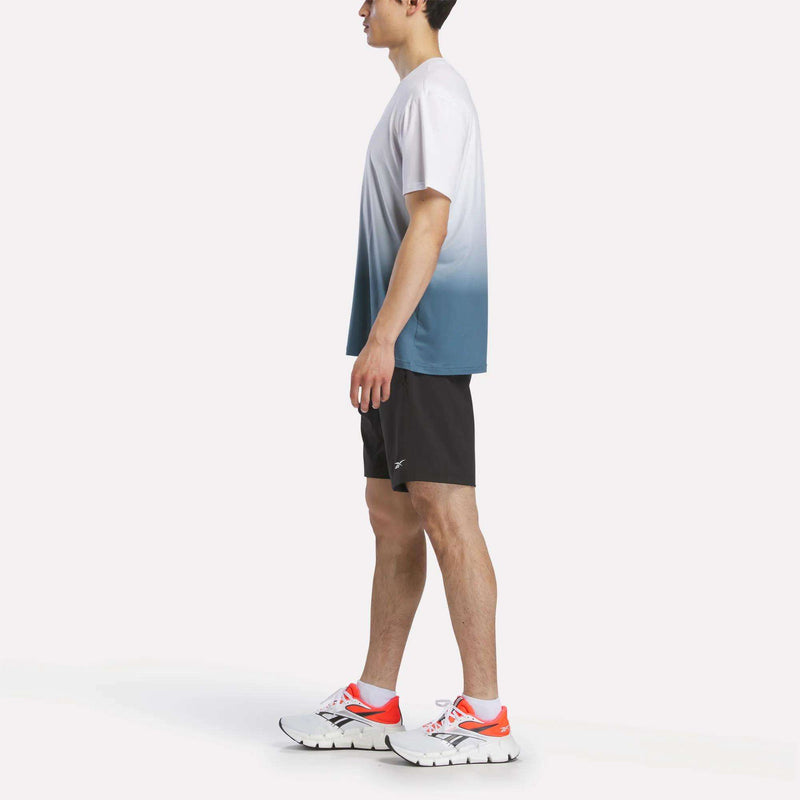 A man stands in profile against a plain white background wearing the Reebok Men's Athlete Speed Ombré T-Shirt in white and blue, paired with black shorts and white running shoes featuring orange accents.