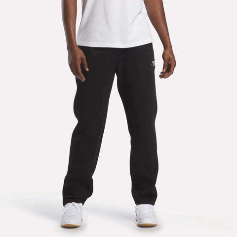 A person stands against a plain white background, with only the lower half of their body visible, showcasing the cozy comfort of the Reebok Men's Fleece Straight Leg Pants in black. They are styled here with a white t-shirt and white sneakers.