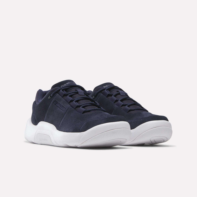 A pair of Reebok DMX Comfort+ Classic walking shoes in navy blue, featuring thick white soles and dark laces, are showcased against a plain light background. Crafted from premium leather, these sneakers boast a sleek, modern design with minimal detailing.