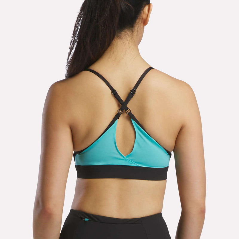 A woman is seen from the back wearing a turquoise Reebok Women's Speed Strappy Sports Bra with black straps and a keyhole cutout, paired with black athletic pants against a plain light background.