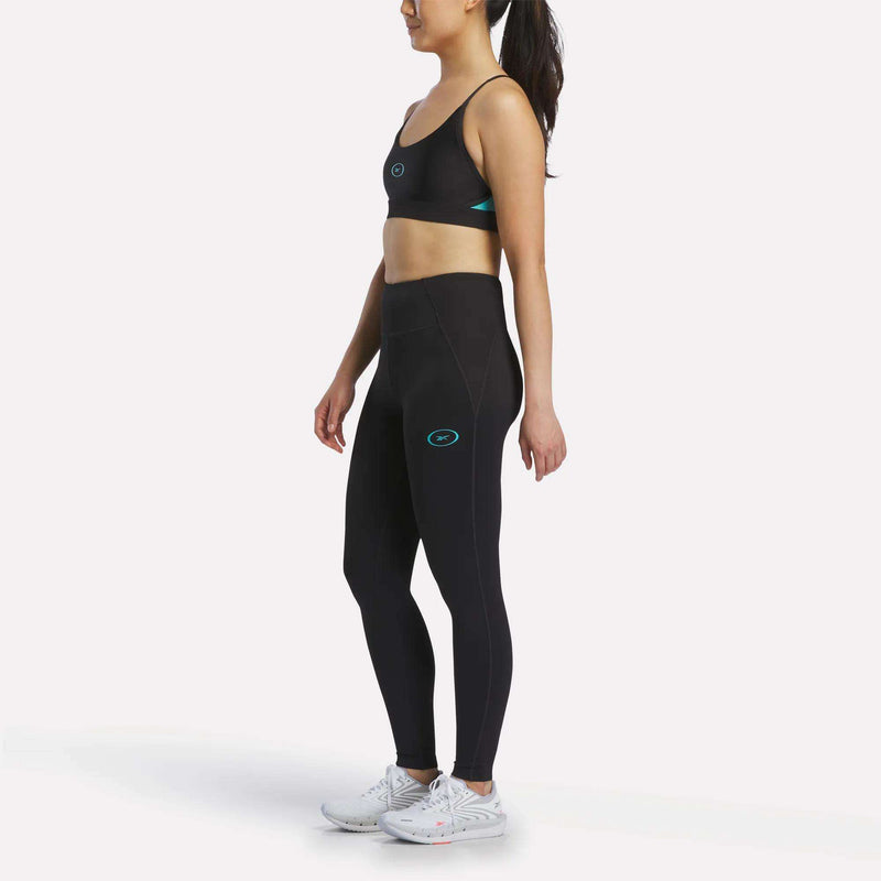 A woman poses in profile wearing the Reebok Women's Speed Strappy Sports Bra, black leggings with teal accents, and white athletic shoes against a plain light background.