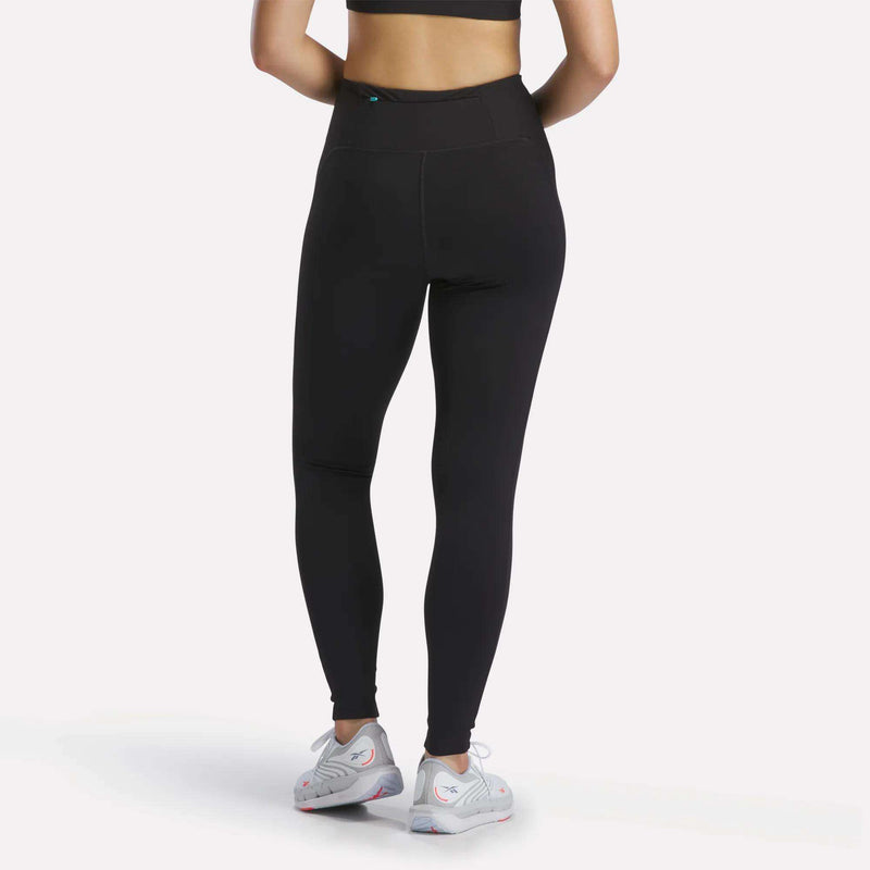A person wearing Reebok Women's Speed HR High-Rise Leggings in black, a black sports bra, and white athletic shoes stands facing away from the camera against a plain white background.
