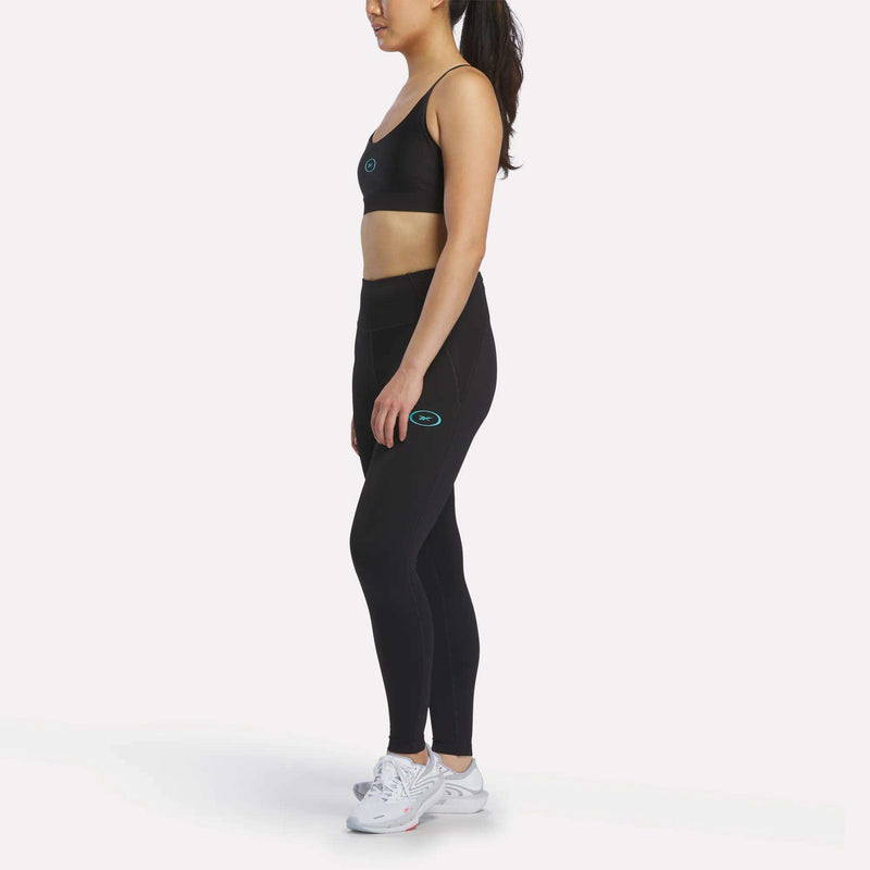 A woman in a black Reebok sports bra and Women's Speed HR High-Rise Leggings stands sideways on a white background, hair in a ponytail. Both clothing pieces feature a small blue and white Reebok logo. She pairs the outfit with white sneakers.