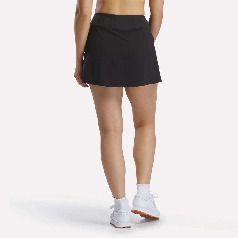 A person stands facing away, wearing the Reebok Women's Athlete Speed Skort 3" in black with built-in shorts, white ruffle-edge socks, and white sneakers against a plain white background.