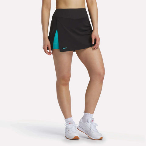 A person wears a black Reebok Women's Athlete Speed Skort 3" with a ribbed waistband, turquoise built-in shorts, white ruffled socks, and white sneakers, standing against a plain white background.