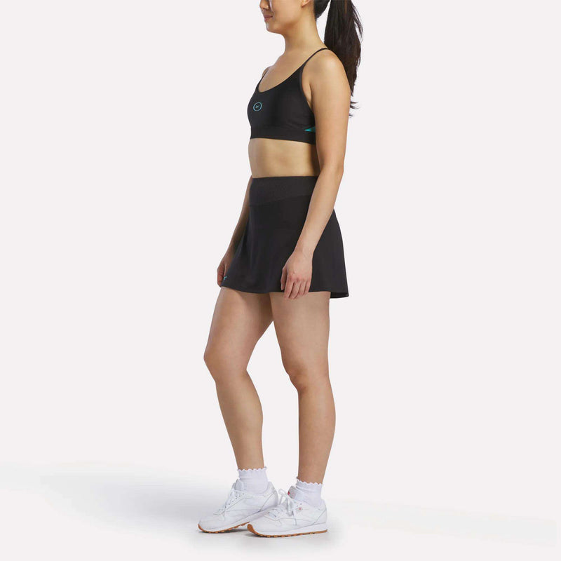 A woman stands in profile wearing a black sports bra, Reebok Women's Athlete Speed Skort 3" with built-in shorts and ribbed waistband, white socks, and white sneakers against a plain white background.