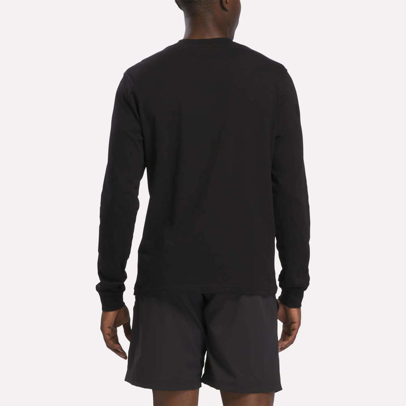 A person is standing facing away, wearing a Reebok Men's Long Sleeve T-Shirt in black and black shorts. The background is plain and light-colored.