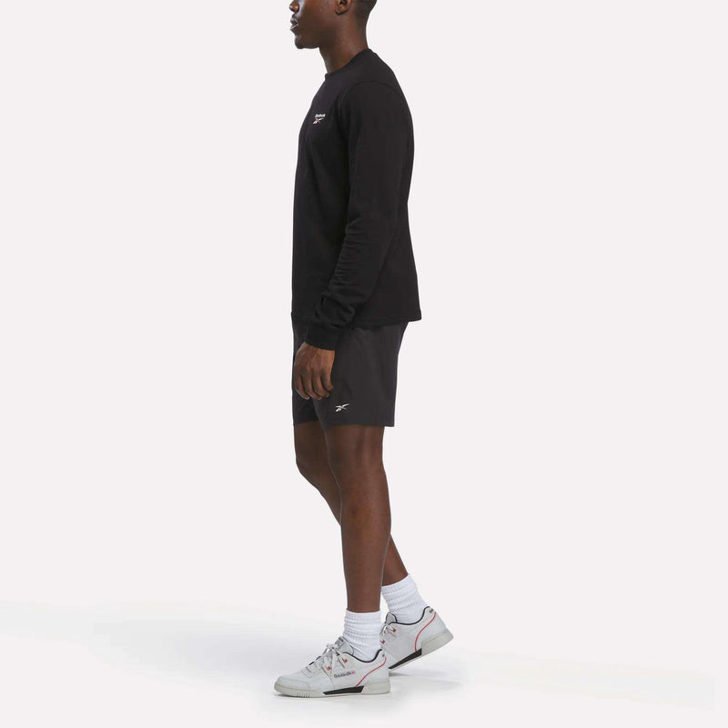 A man in a black Reebok Men's Long Sleeve T-Shirt, black shorts, white socks, and white Reebok sneakers stands facing left against a plain white background.