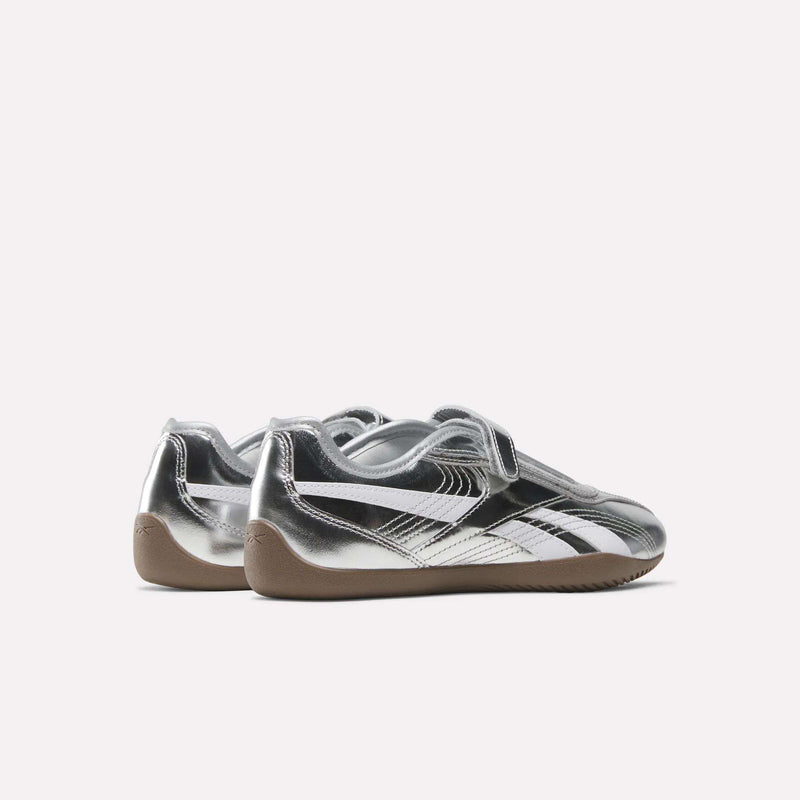 A pair of Women’s Ultra Lo Mary Jane Shoes by Reebok, featuring a metallic silver finish with black and white accents, Velcro straps, and gum rubber soles, are displayed on a white background.
