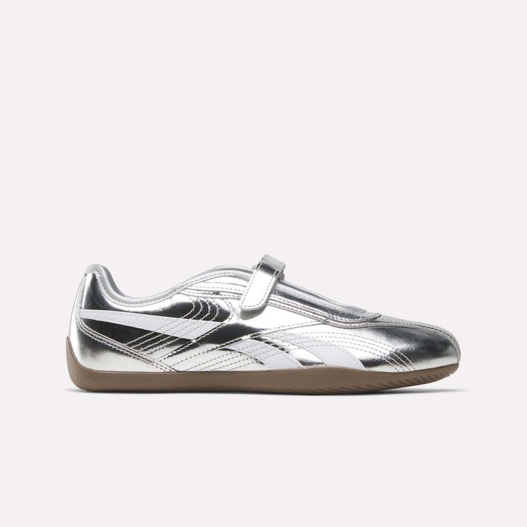 The Reebok Women’s Ultra Lo Mary Jane Shoes in Silver/White/Gum showcase a classic Mary Jane-inspired silhouette with a metallic silver upper, Velcro strap, white and black detailing, and a light brown gum sole, presented in a low-profile side view against a plain light background.
